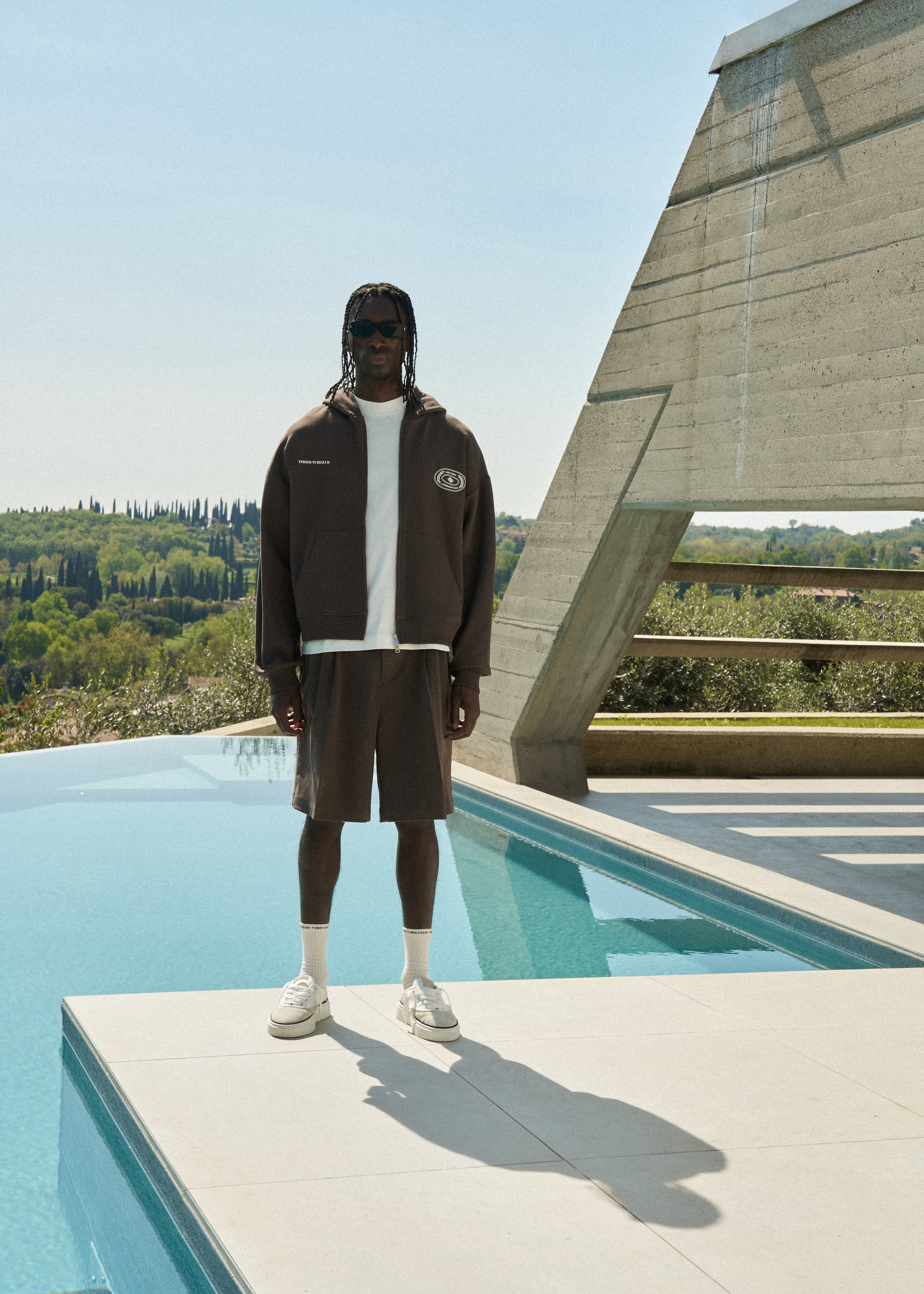 Man standing by a pool in a brown hoodie and brown shorts with a modern architectural structure in the background