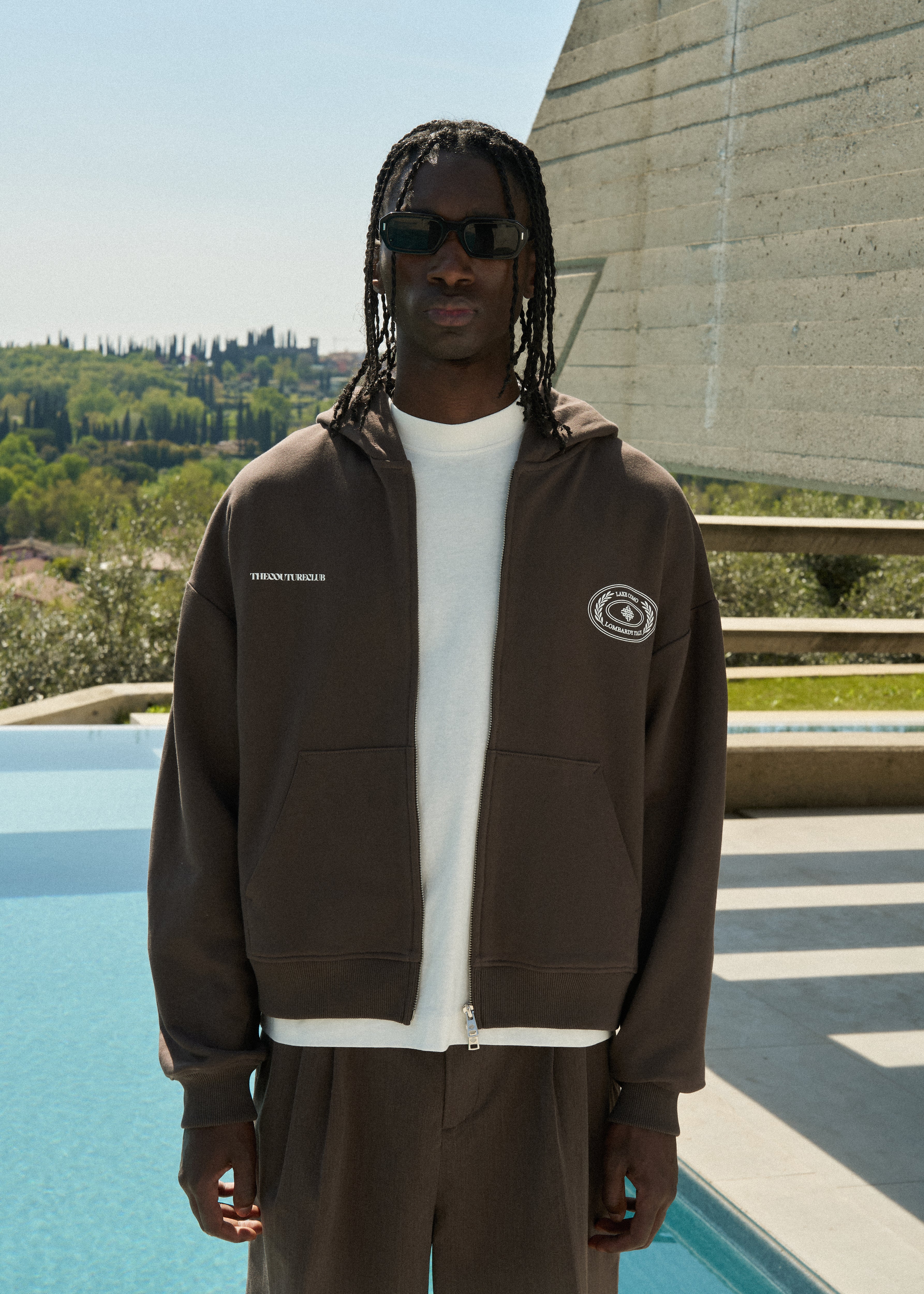 Man wearing a brown hoodie with a logo, standing by a pool with a scenic background.