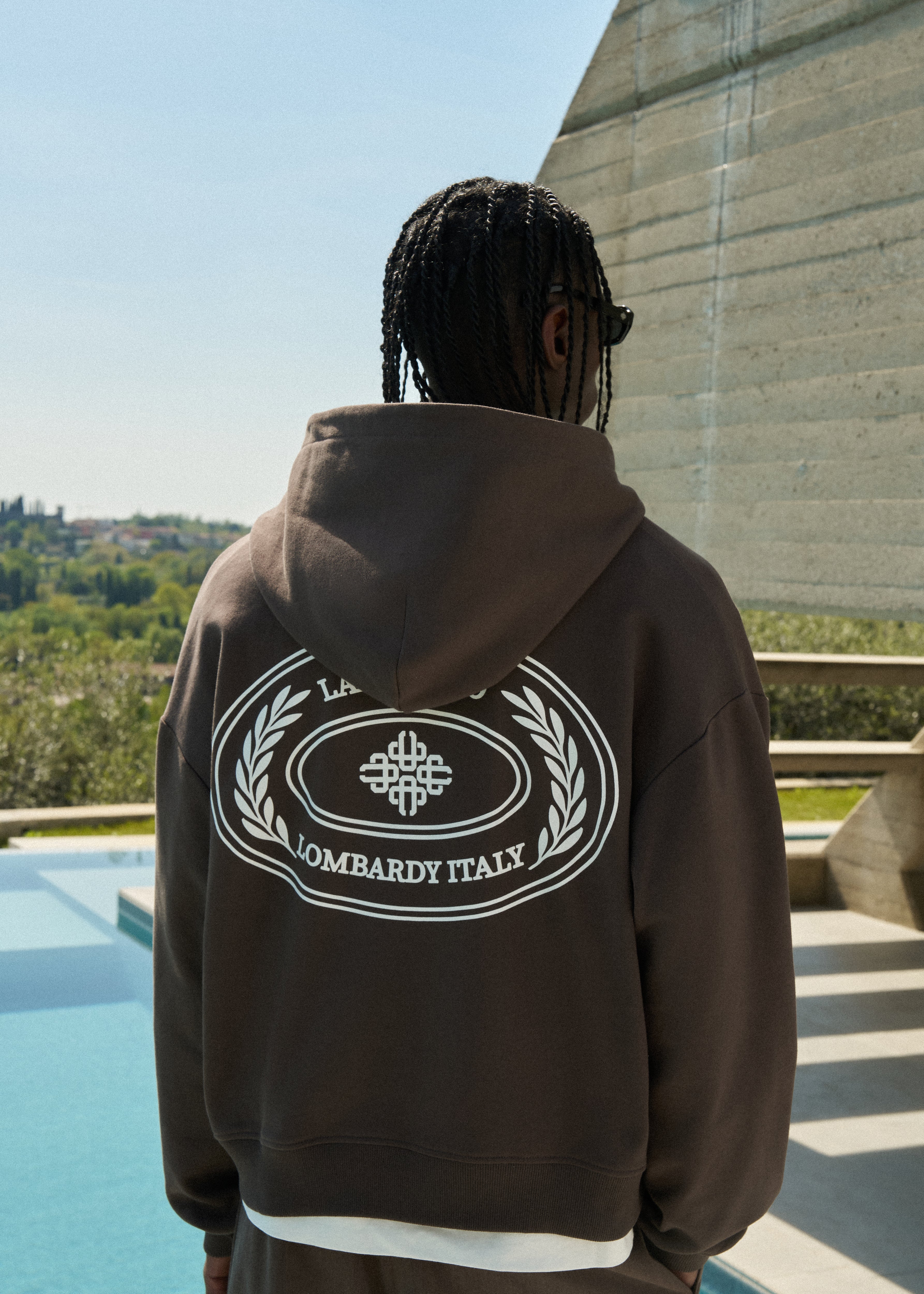 Man wearing a brown hoodie with a logo and text on the back, standing by a pool with a scenic background.