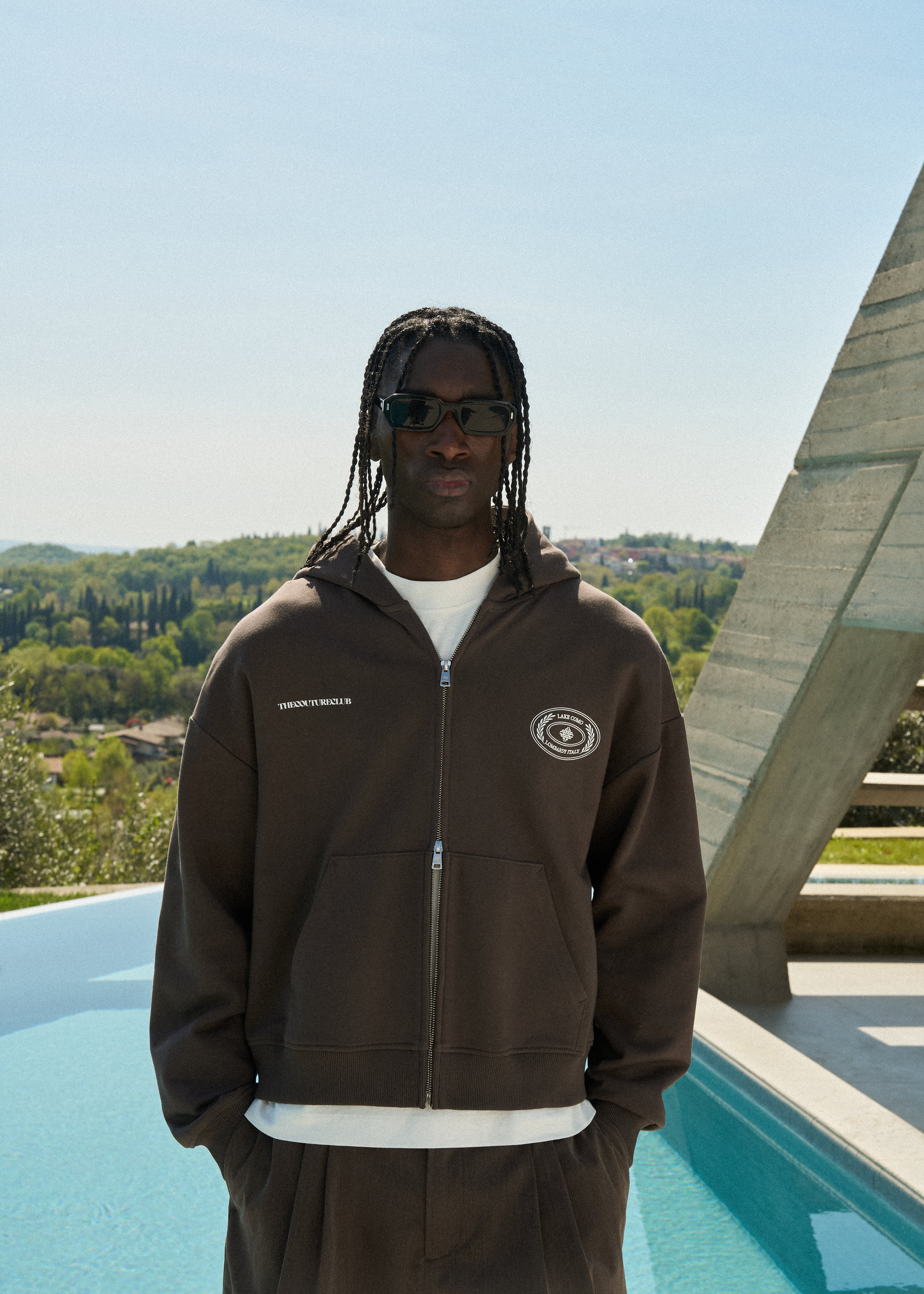Man wearing a brown hoodie with a logo, standing by a pool with a scenic background.