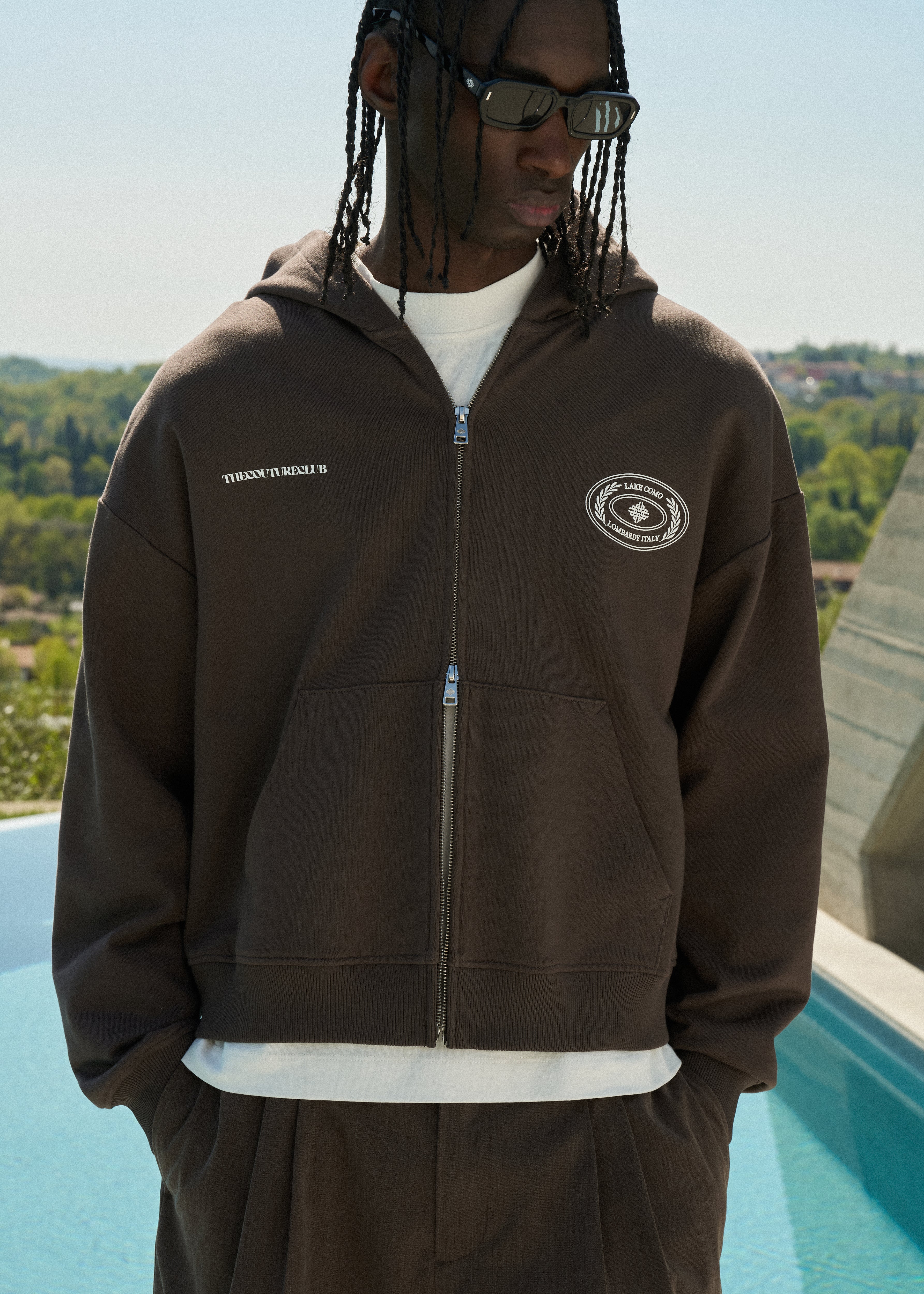 Man wearing a brown hoodie with logos, standing by a pool with a scenic background.