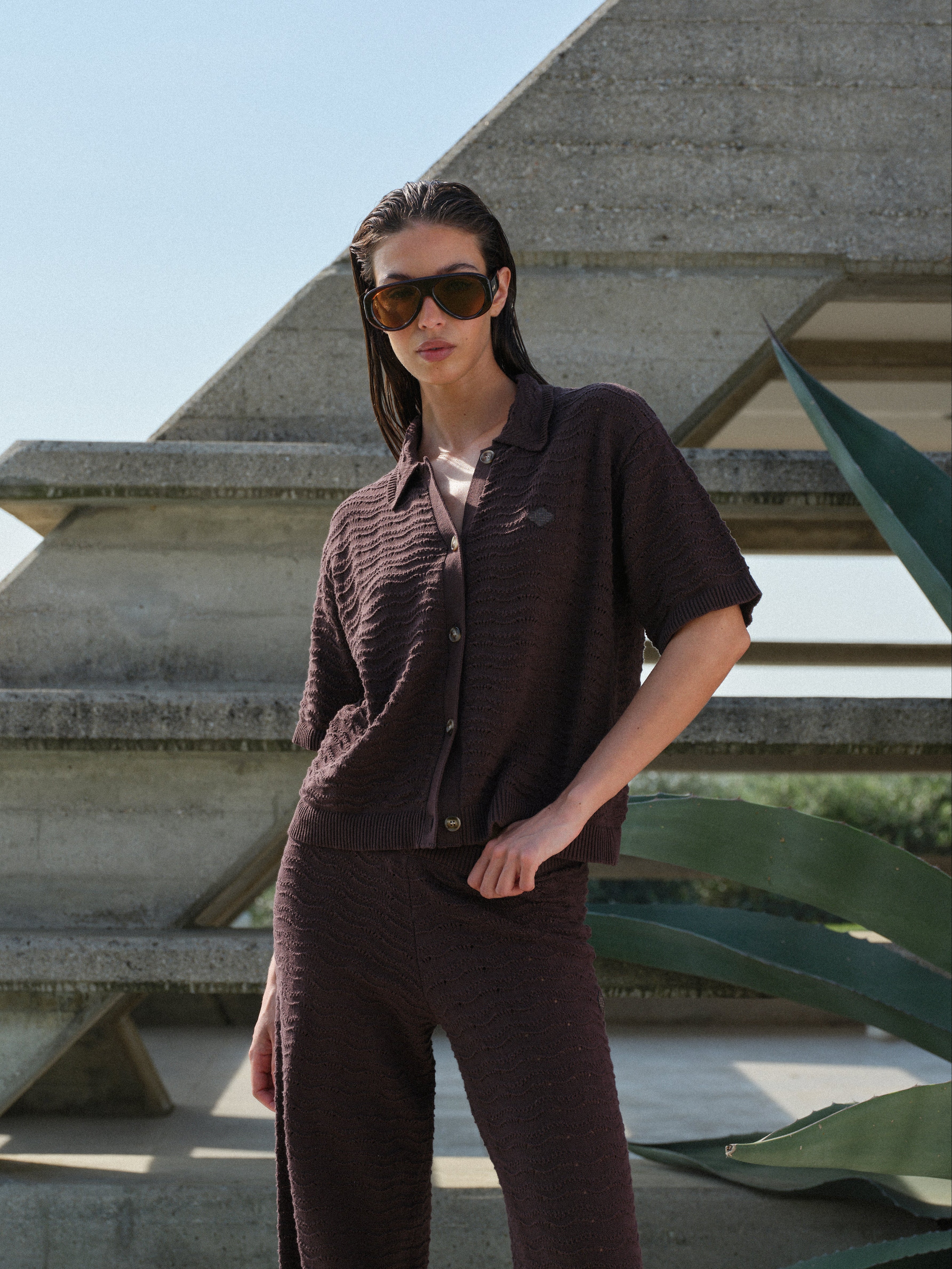 Woman in a brown crochet knit shirt with matching pants standing in front of a geometric architectural structure.