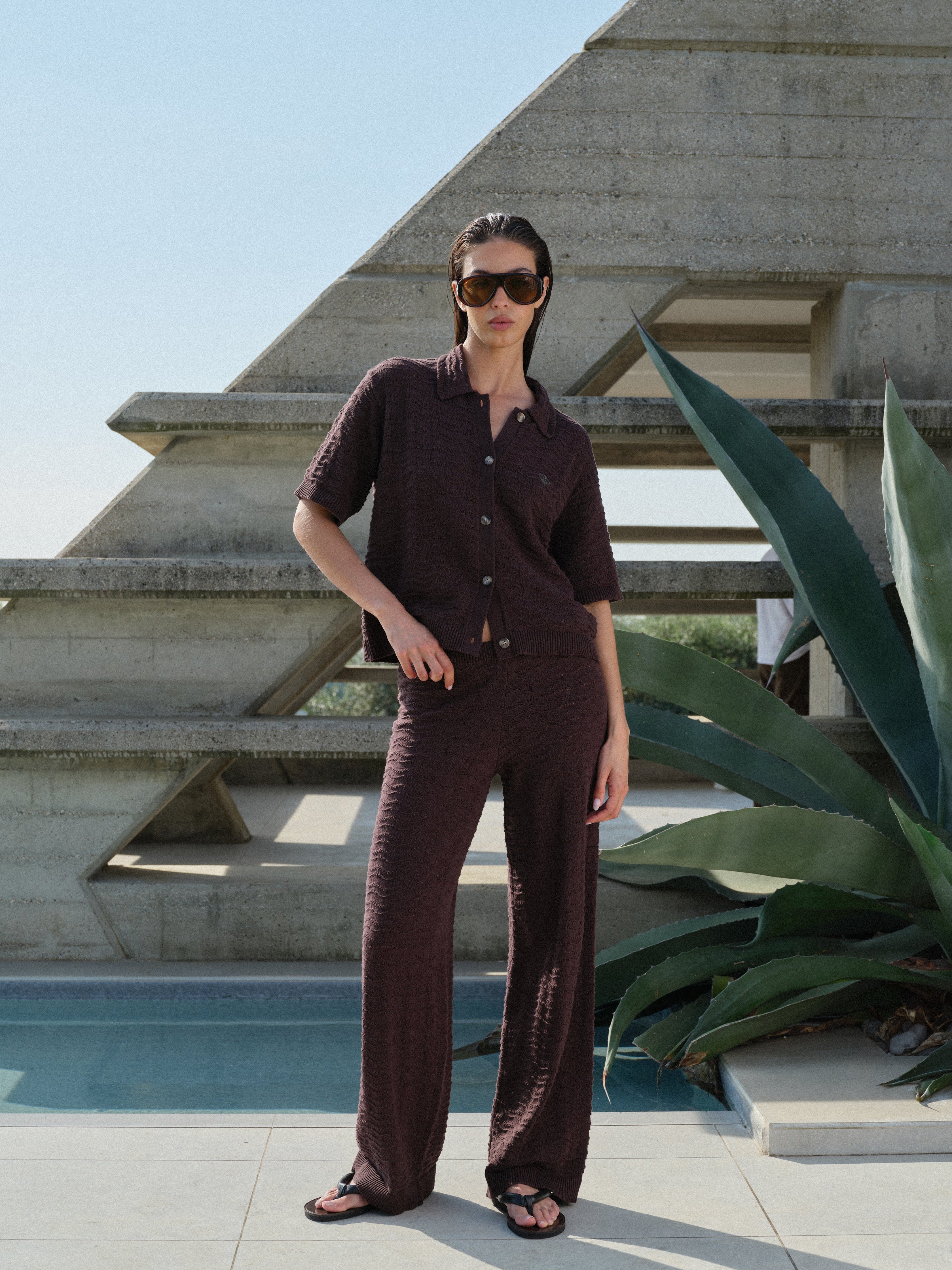 Woman in a dark brown crochet knitted matching set standing by a pool with a modern building and plant in the background