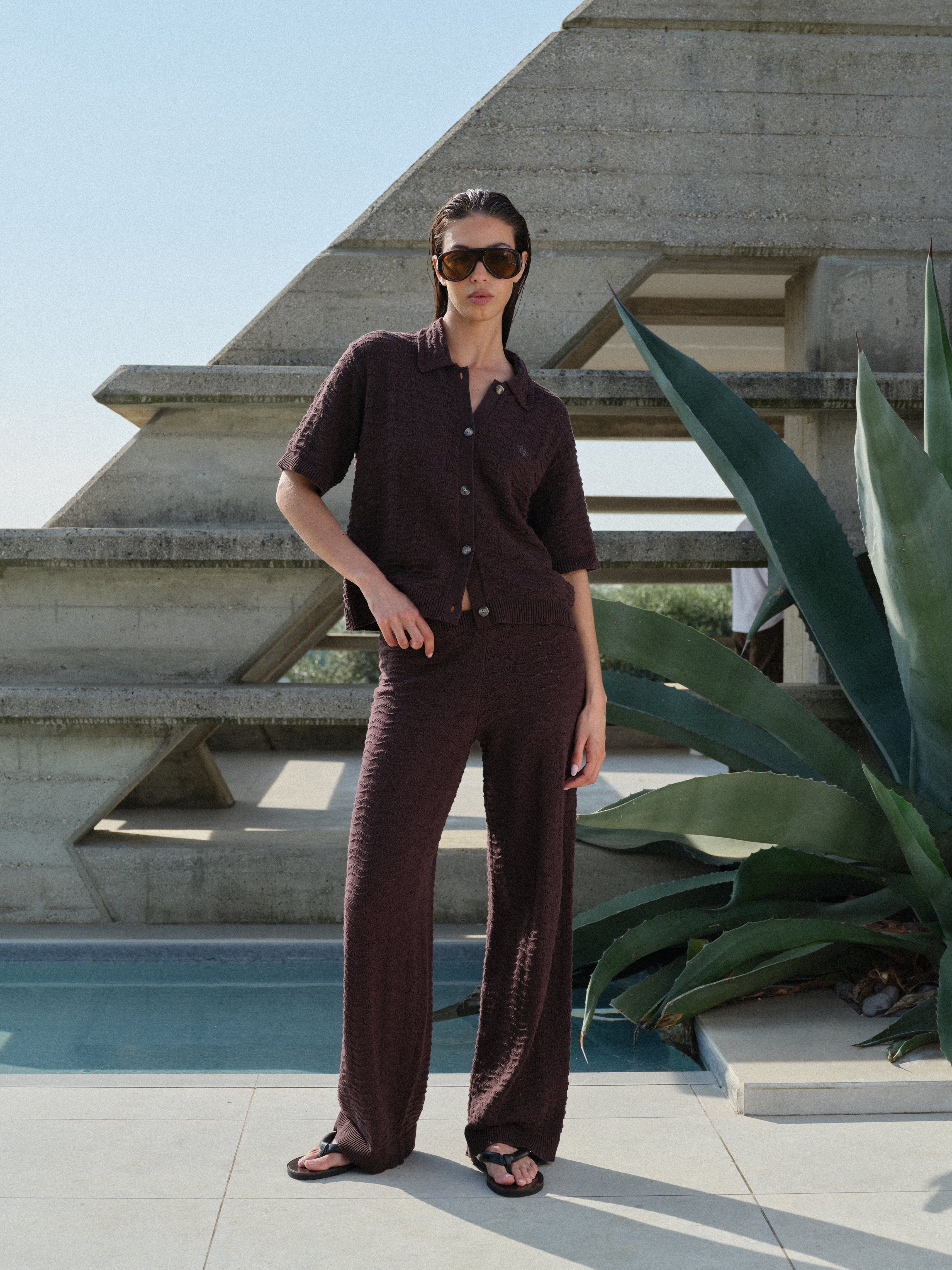 Woman in a brown crochet knit shirt with matching pants and sunglasses standing in front of a geometric architectural structure.