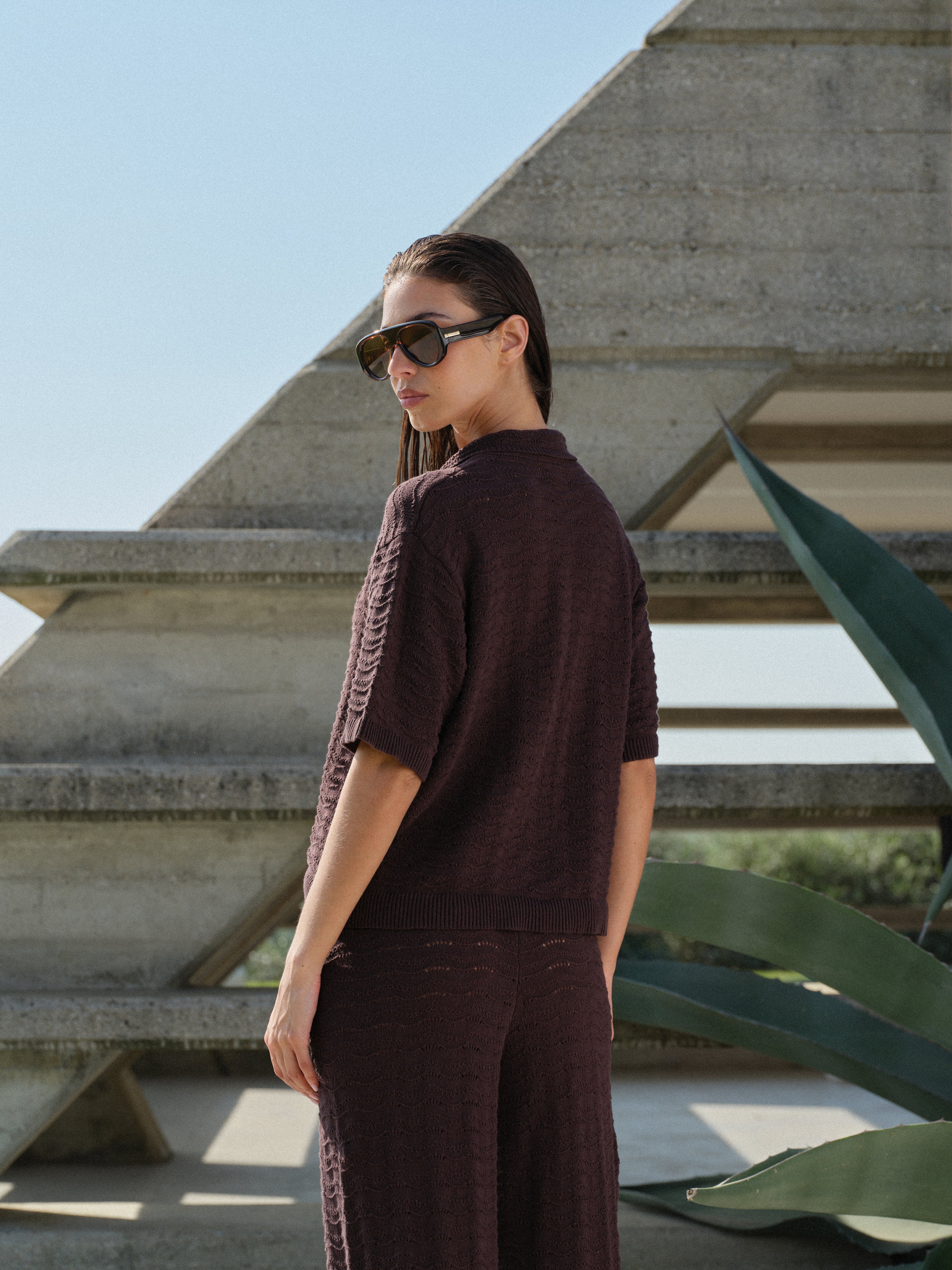 Woman in a brown crochet knit shirt with matching pants and sunglasses standing in front of a geometric architectural structure.