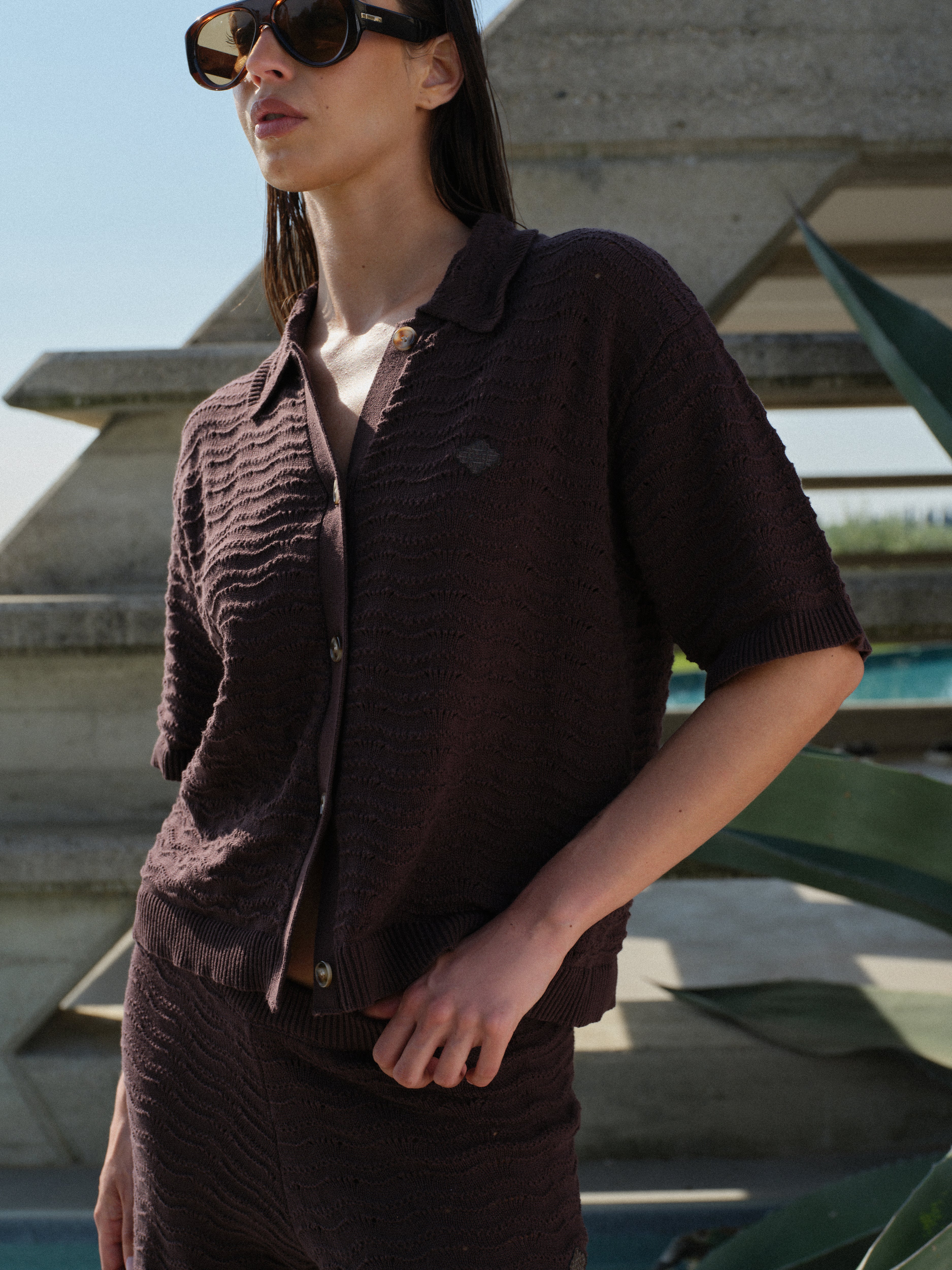 Woman in a brown crochet knit shirt with matching pants and sunglasses standing in front of a geometric architectural structure.