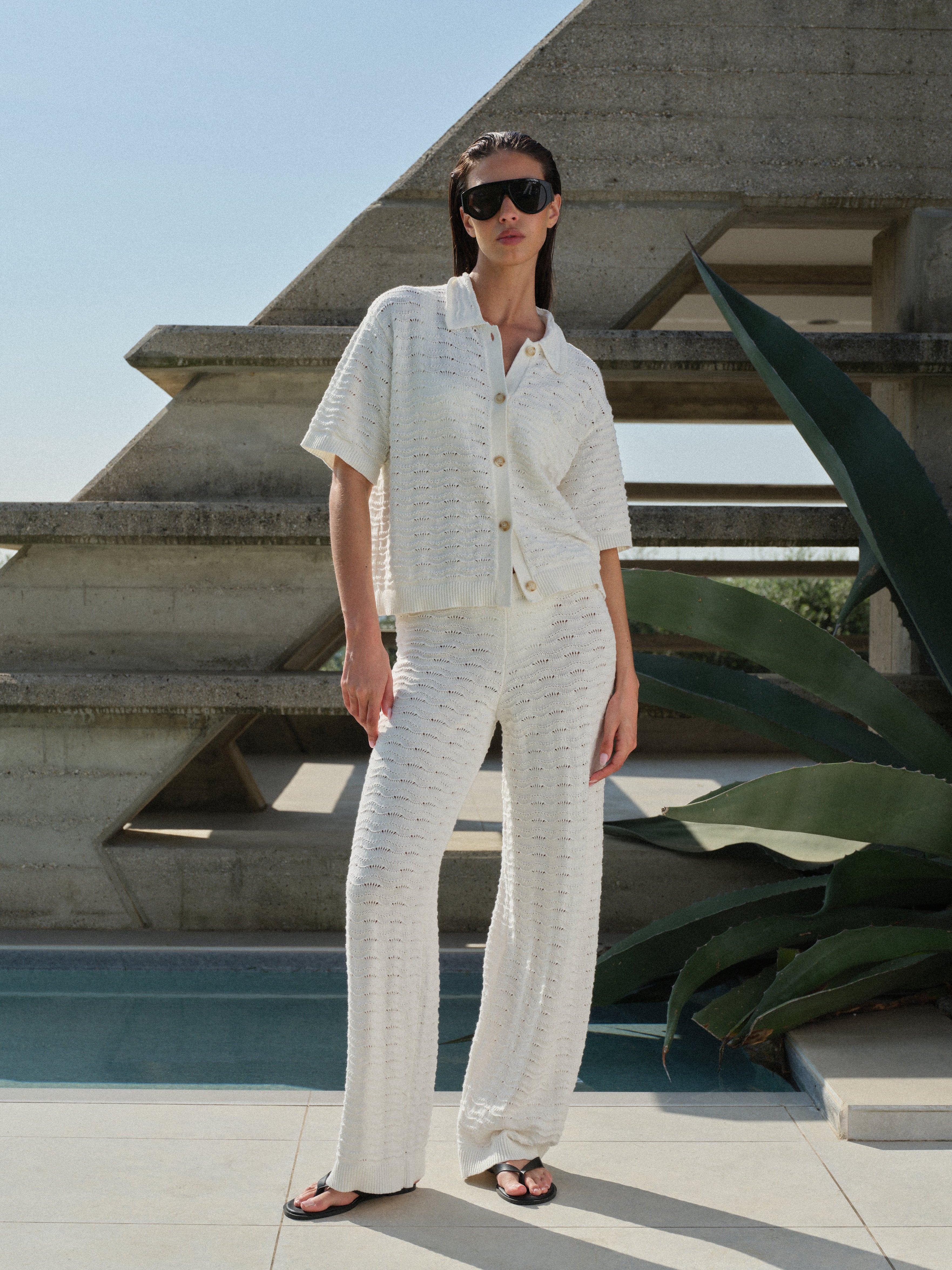Woman in a white knit crochet shirt, sunglasses and matching pants standing outdoors with a concrete structure and greenery in the background.