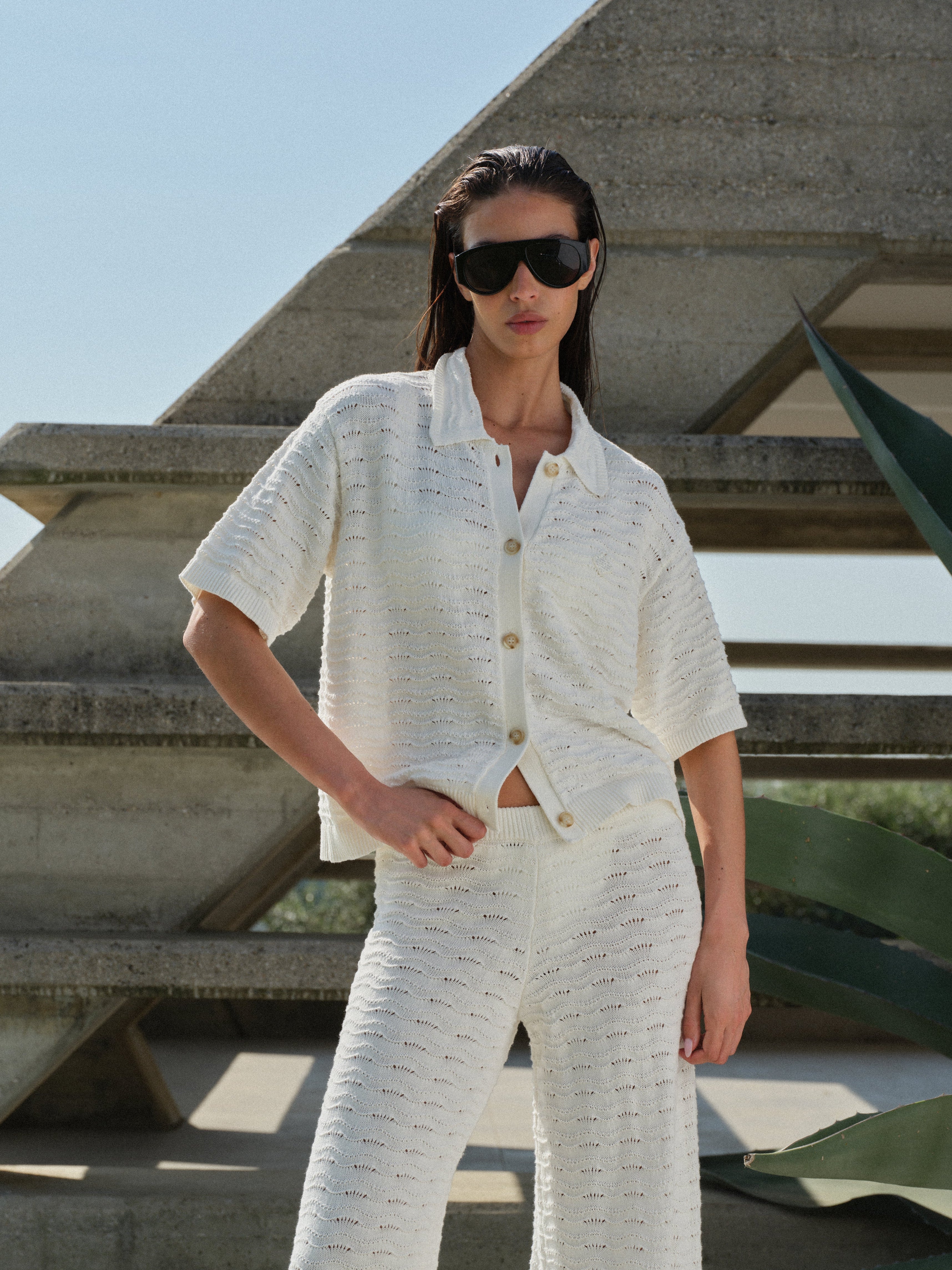 Woman in a white knit crochet shirt, sunglasses and matching pants standing outdoors with a concrete structure and greenery in the background.