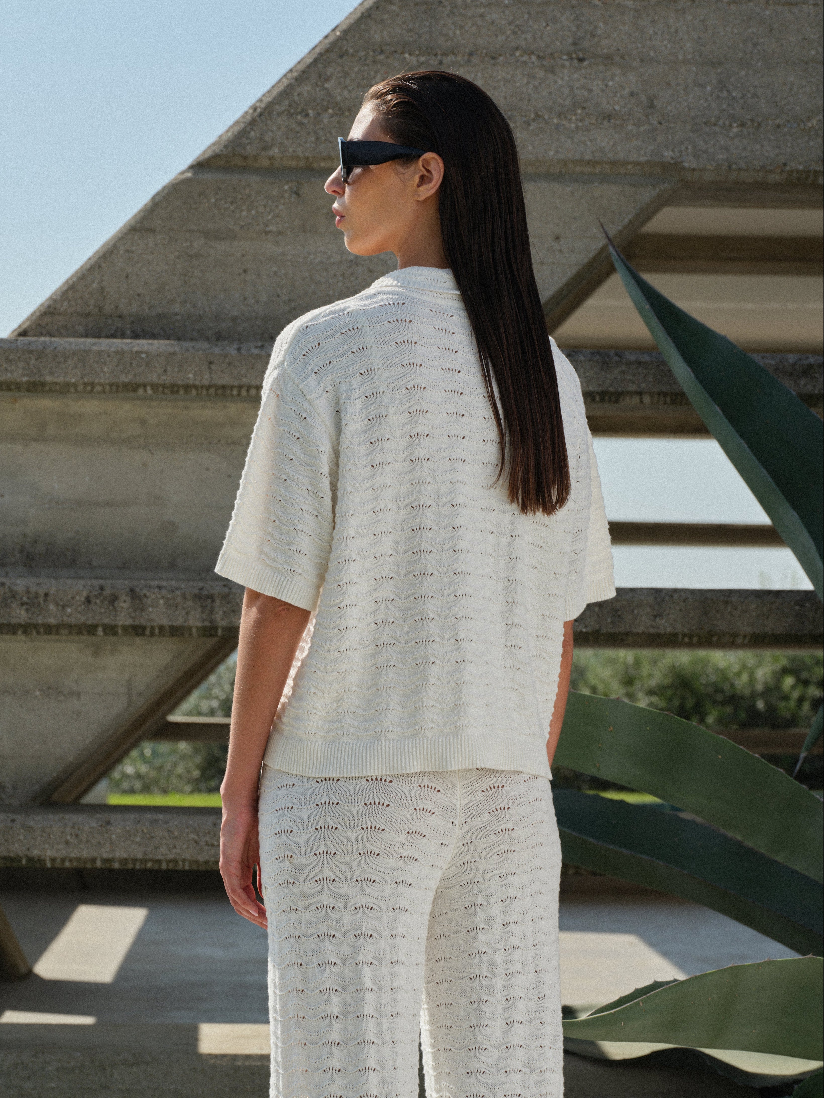Woman in a white crochet knit matching set standing in front of a modern architectural structure.