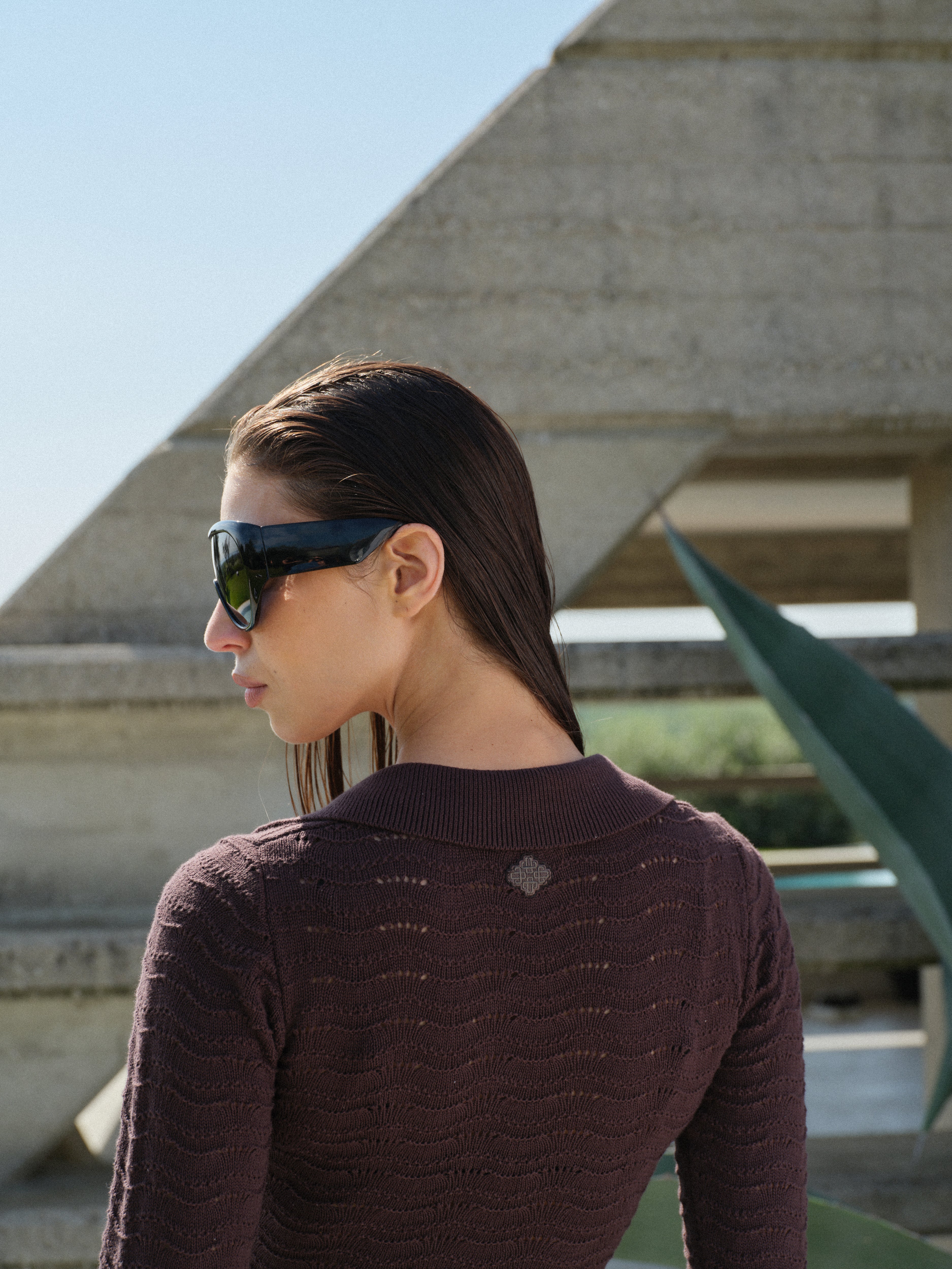Woman wearing a brown long-sleeved crochet knitted dress and sunglasses standing in front of a modern architectural structure.