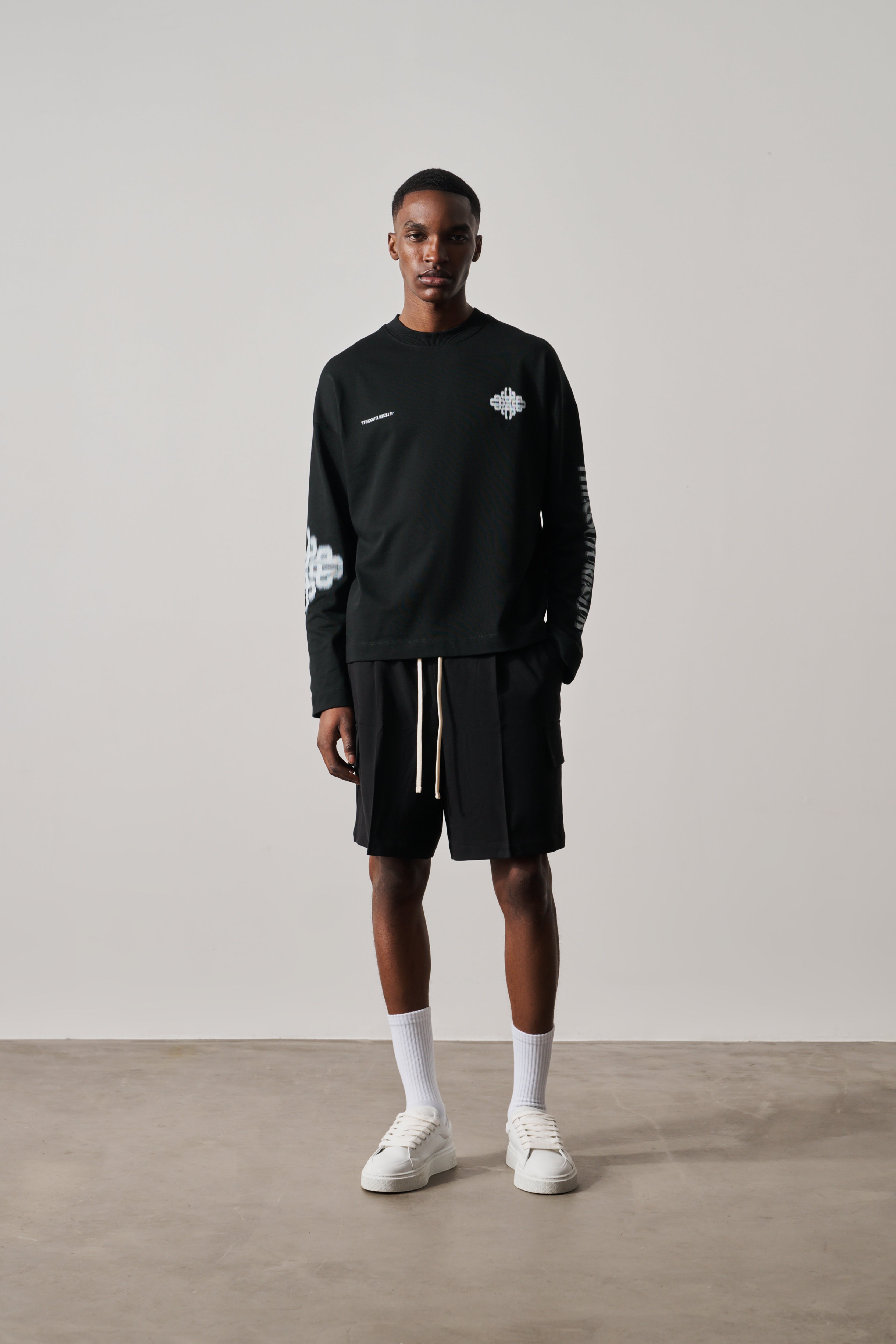 Person wearing a black long-sleeve t-shirt and shorts with white sneakers on a plain background