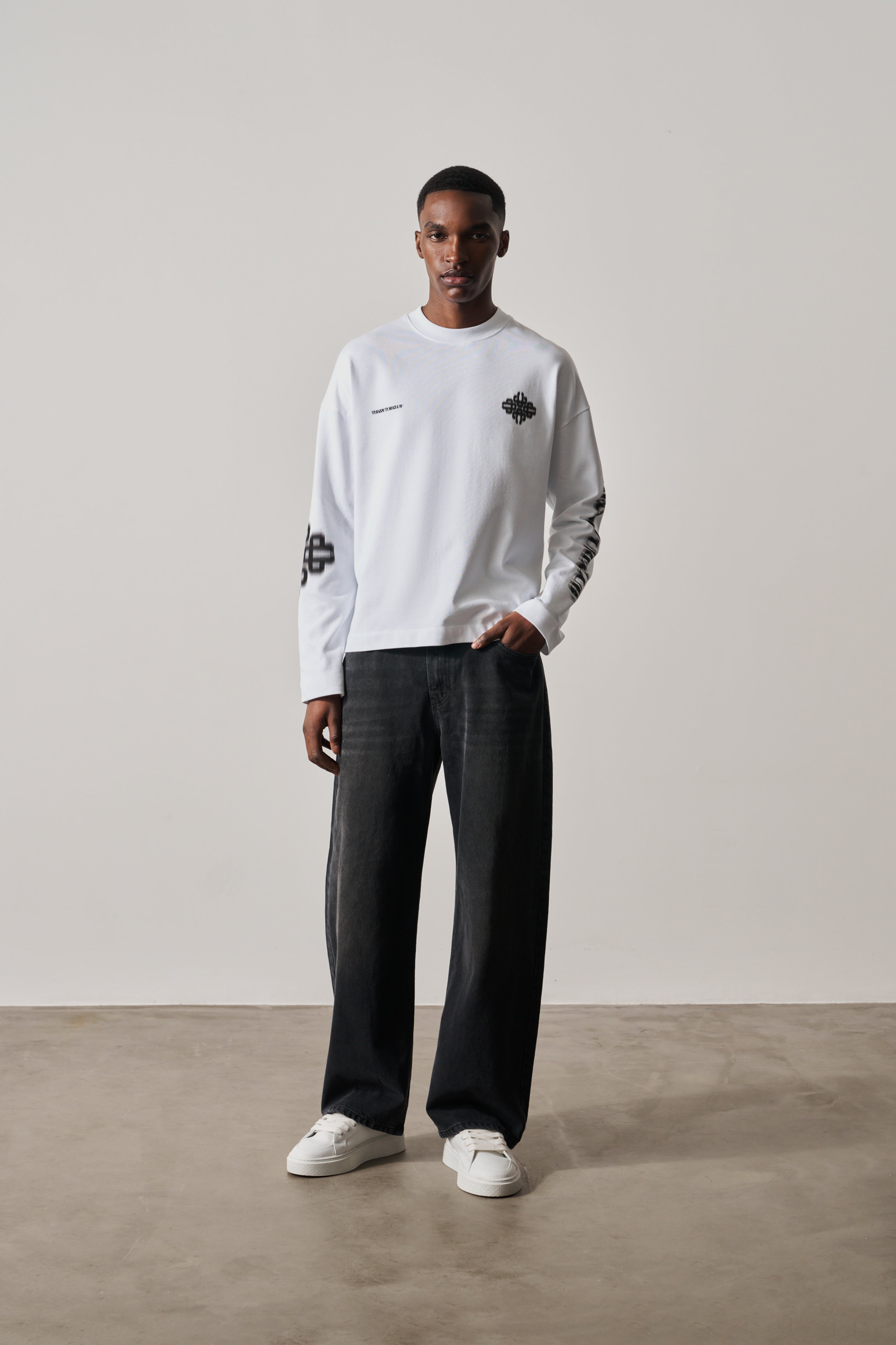 Person wearing a white long-sleeve t-shirt with black graphics and dark pants on a plain background