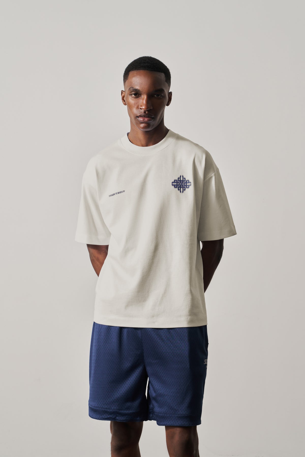 Man wearing a beige t-shirt with a logo and blue shorts on a light gray background